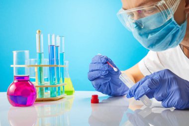 Genç bayan teknisyen ya da bilim adamı sıvı örneğini pipetle şişeye dolduruyor. Mavi arka planda çiçekli test tüpleri.