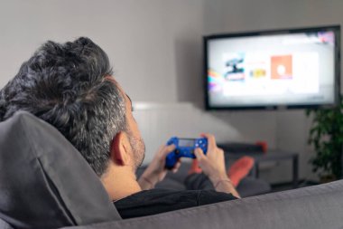 Evde video oyunu oynayan bir adam elinde oyun konsolu ve joystick ile televizyon karşısında koltukta oturuyor.