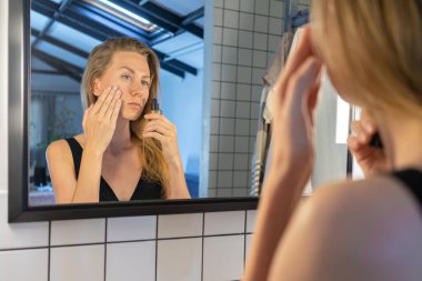 Güzel bir kadın yüz nemlendirici sürüyor, krem sürüyor, banyo aynasının önünde duruyor. güzellik tedavisi