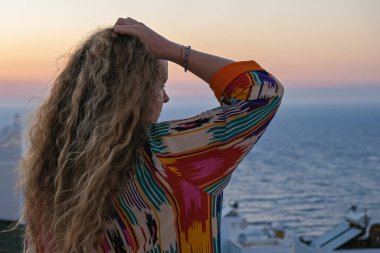 Balkonda bir kadın güzel, renkli günbatımına bakıyor ve elini saçlarında görüyor.