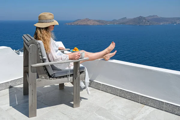 Hermosa mujer joven sombrero de paja y taza de café sentado en el balcón terraza blanca de la casa o el hotel con vista al mar —  Fotos de Stock