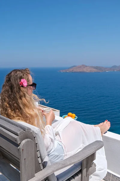 Hermosa mujer joven con flor en taza de café de pelo sentado en el balcón de la terraza blanca de la casa o el hotel con vista al mar —  Fotos de Stock