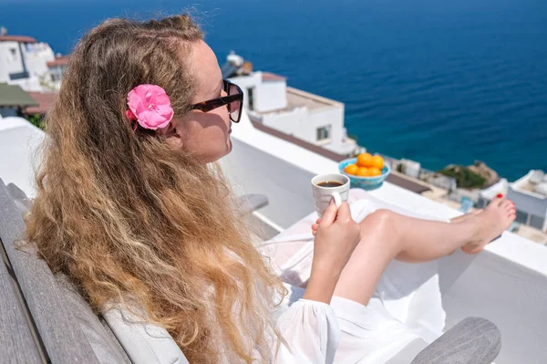 Hermosa mujer joven con flor en taza de café de pelo sentado en el balcón de la terraza blanca de la casa o el hotel con vista al mar —  Fotos de Stock