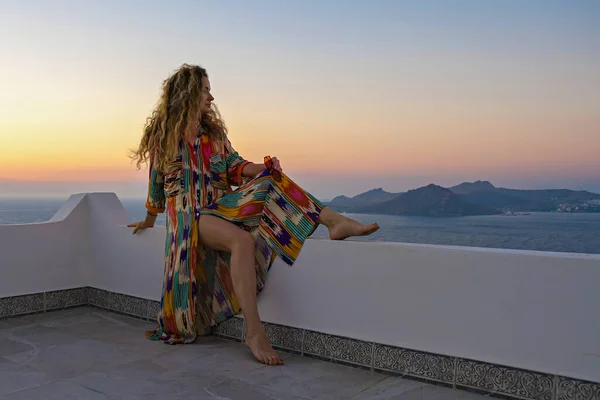 Gyönyörű nő színes ruhában, erkélyes teraszon, kilátással a tengerre és a naplementére. üdülőhely. nyári szabadság koncepciója — Stock Fotó
