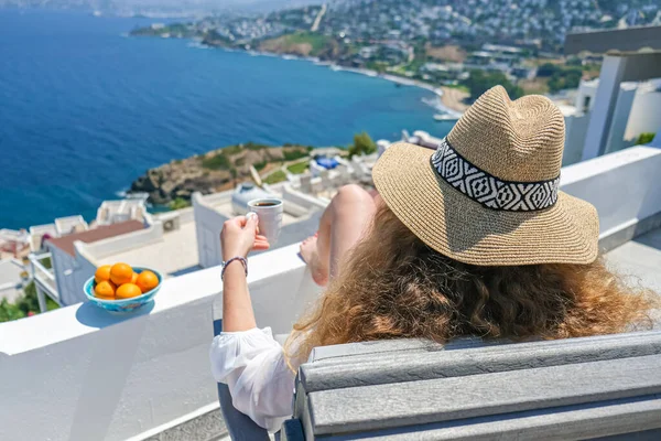 Hermosa mujer joven sombrero de paja y taza de café sentado en el balcón terraza blanca de la casa o el hotel con vista al mar —  Fotos de Stock