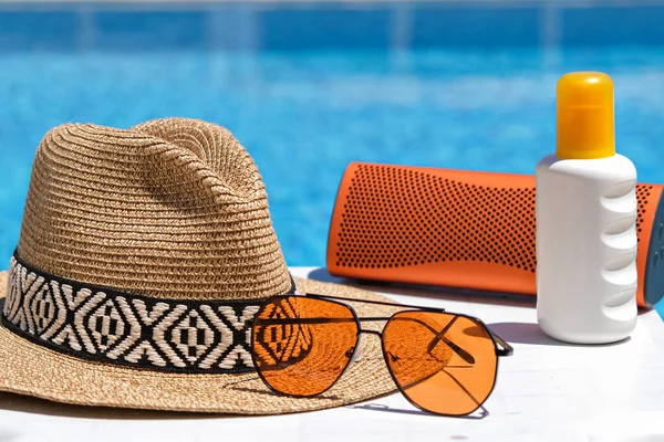 orange coloured beach accessories near swimming pool. Sun cream, sunglasses, music speaker and straw hat.