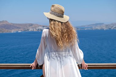 Beyaz mayo ve hasır şapkalı güzel bir kadın. Deniz manzaralı beyaz teras balkonunda..