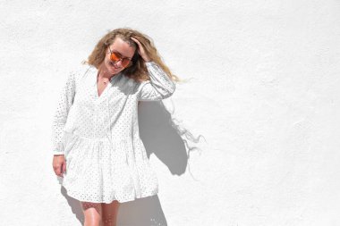 Beyaz elbiseli, turuncu gözlüklü, beyaz taş duvarlı güzel, mutlu bir kadın. Arka plan fotokopi alanı..