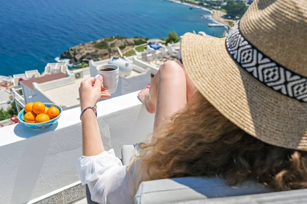 Beyaz elbiseli, hasır şapkalı ve kahve fincanlı güzel bir kadın. Deniz manzaralı beyaz teras balkonunda oturuyor..