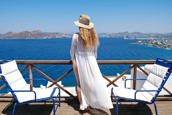 Gyönyörű nő hátulról fürdőruhában fehér ruha és szalma kalap fehér terasz erkéllyel ház vagy szálloda kilátással a tengerre — Stock Fotó