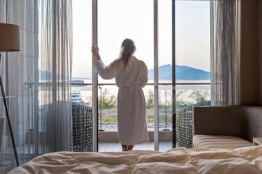Bornozlu güzel bir kadın otel odasının terasında duruyor. Deniz manzaralı sabah güneşiyle birlikte..