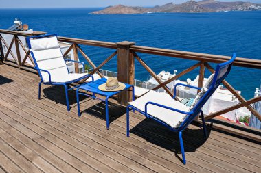 İki beyaz sandalyesi, masası ve deniz manzaralı ahşap bir villa ya da otel terası.