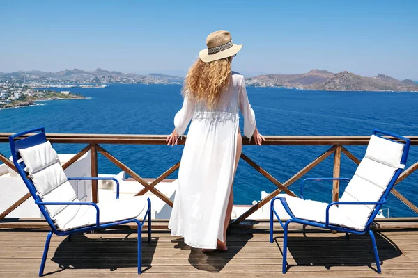 Bella donna da dietro in costume da bagno vestito bianco e cappello di paglia sulla terrazza bianca balcone di casa o hotel con vista mare — Foto Stock