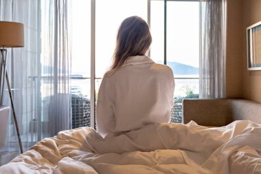 Sabahleyin bornozlu bir kadın yatağında oturmuş, pencereye bakıyor ve otel manzarası seyrediyor.