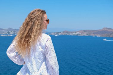 Beyaz elbiseli, güneş gözlüklü güzel bir kadın villa otelinin beyaz terasında Sea View 'a bakıyor.