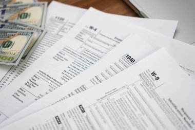 Çeşitli vergi formlarına konulan dolar banknotları, yasalara uygun olarak ve yardım hizmetine ihtiyaç duyulan karmaşık belge doldurma sürecine işaret etmektedir.