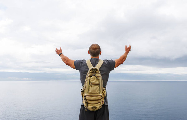 Traveler with backpack stand on the shore and looking at the sea with raised hands in air. Rear view