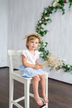 Genç bir sıcak gülümseyen kız beyaz sandalyeyle çiçek çelenk ile duvarın yanında oturuyor. Dikey portre