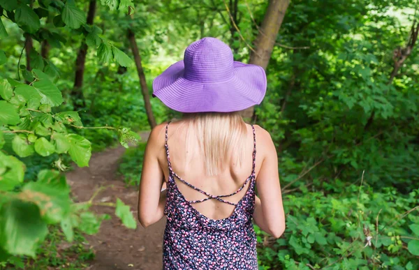 A woman in a violet hat and in violet dress walks through the woods. View from the back.