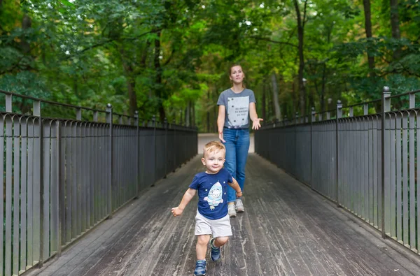 Happy young mother and her running little son on the bridge in public park. Selective focus in kid