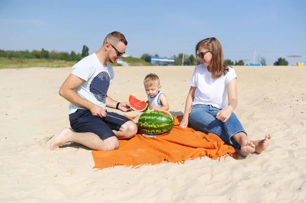 若い家族は 湖の砂浜でスイカを一緒に食べています お互いを探してください — ストック写真