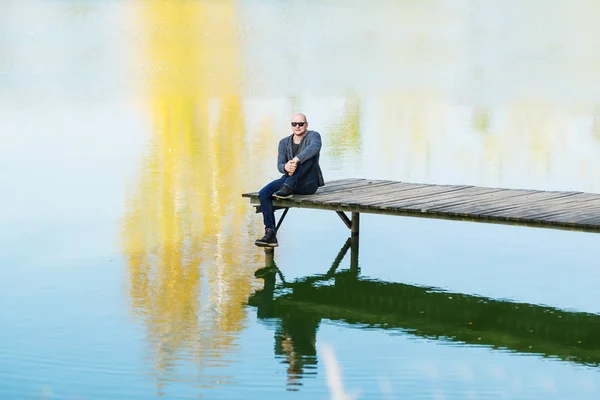 Lysy Młody Człowiek Czarne Okulary Przeciwsłoneczne Siedzieć Molo Pobliżu Jeziora — Zdjęcie stockowe