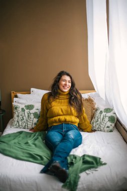 Güzel genç gülümseyen esmer kot pantolon ve kazak ve uzak yatakta otururken arıyor