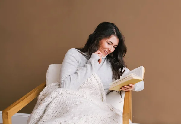 Mujer joven pasando un día relajante en casa sentada en un sillón leyendo un libro con una sonrisa feliz — Foto de Stock