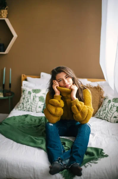 Позитивна жінка в теплому светрі сидить у ліжку і посміхається до камери вдома — стокове фото
