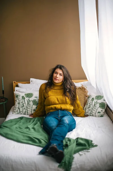 Портрет красивой юной брюнетки в джинсах и свитере, сидящей на кровати — стоковое фото