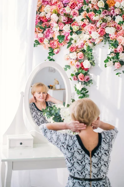 Beautiful senior woman with blonde hair looking at reflection in mirror in white studio with colorful flowers