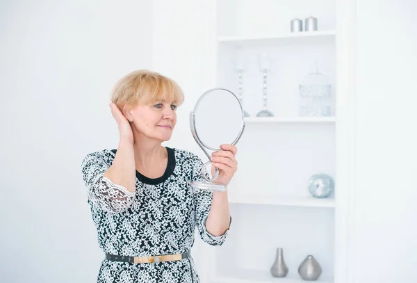 Una hermosa mujer mayor de pie en una habitación blanca y mira en el pequeño espejo — Foto de Stock