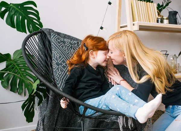 Мама целует маленькую рыжую дочь, сидя в кресле в интерьере дома . — стоковое фото