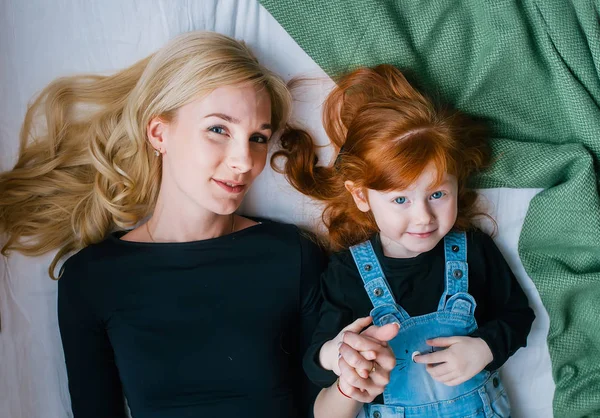 Вид сверху на красивую молодую мать и ее маленькую рыжеволосую дочь, глядя в камеру, лежа дома в постели — стоковое фото
