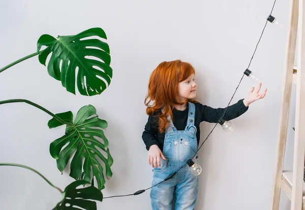 Bastante pelirroja niña pequeña con grandes hojas verdes y una guirnalda ligera . — Foto de Stock