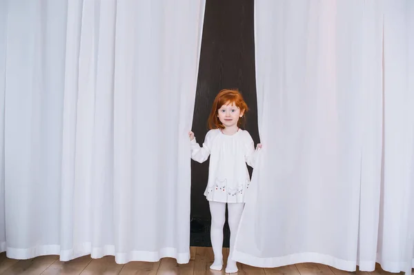 Menina ruiva em pé no chão na sala com cortinas brancas . — Fotografia de Stock