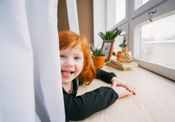 Pretty redhead litlle girl sitting at table near window and she puts out the tongue — Stock Photo, Image