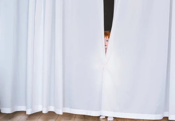 A pequena criança ruiva olha para fora por causa de cortinas brancas — Fotografia de Stock