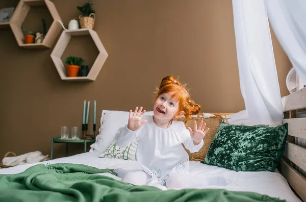 Criança ruiva engraçada sentada na cama em casa. Ele tem um sorriso engraçado na cara. . — Fotografia de Stock