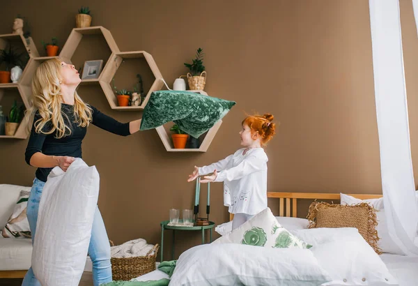 Щаслива молода мати сміється і грає на подушці битися зі своєю маленькою рудою дочкою на ліжку . — стокове фото