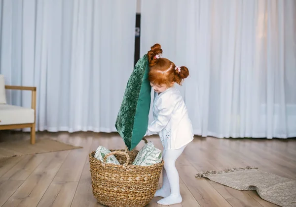 Feliz pelirroja niña en pijama jugar con la cesta y almohadas en el suelo en casa . — Foto de Stock