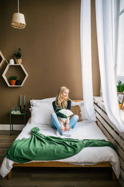 Блондинка мріє в ліжку біля світлого вікна вдома — стокове фото