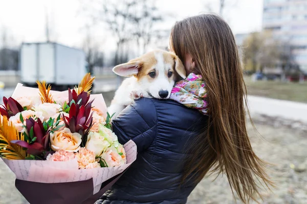 Mujer joven y bonita con ramo de flores sosteniendo perro pequeño, sobre el fondo de la calle de la ciudad. Vista trasera — Foto de Stock