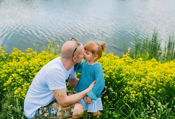 Un padre caucásico sosteniendo y besando a una niña antes de un lago en un prado . —  Fotos de Stock