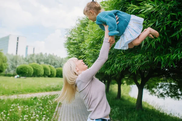 A young mother plays with her little daughter in the park, hold on to her hands. Throw the girl in the air. — Stock Photo, Image