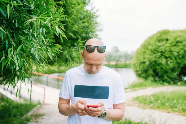 Φαλακρός άνθρωπος με γυαλιά ηλίου κρατώντας το τηλέφωνο σε ένα πάρκο — Φωτογραφία Αρχείου