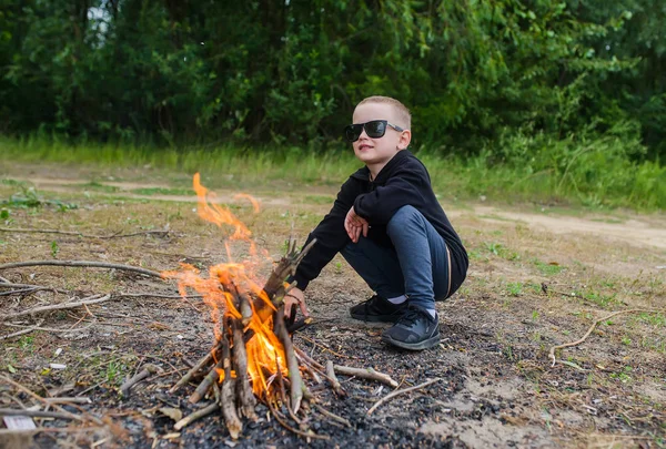 Cute Boy w okulary sprawia, że ogień w lesie do gotowania. Kemping z dziećmi w letnim lesie. — Zdjęcie stockowe