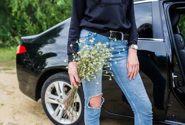 Woman in jeans with a beautiful bouquet of wildflowers against the black car on summer day — Stock Photo, Image