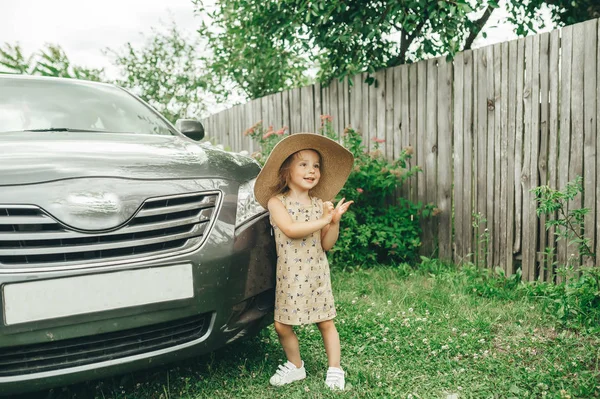 Carino bambina in cappello di paglia in piedi vicino auto nel villaggio — Foto Stock