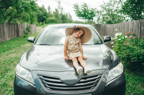 Carino bambina in cappello di paglia seduto sul cappuccio auto in cortile — Foto Stock